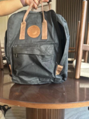 Svart ryggsäck - Klassisk svart ryggsäck med bruna detaljer i läder på handtag. Rymligt huvudfack, framficka med dragkedja och slitstarkt material. Perfekt för skola eller utflykter, enkel och stilren design. Finns fåtal små fläckar. Köpes i befintligt skick.