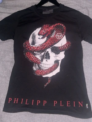 Svart Philipp Plein t-shirt med orm - Svart t-shirt från Philipp Plein med stor vit dödskalle och röd orm på framsidan samt röd text på både fram- och baksida. T-shirten har korta ärmar och är tillverkad i bomull. Snygg och edgy design som sticker ut.