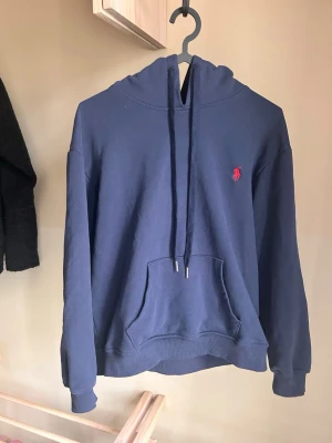 Blå hoodie från Polo Ralph Lauren - Jag är 165. Snygg blå hoodie från Polo Ralph Lauren med klassisk röd logga på bröstet. Hoodien har en stor känguruficka, ribbade muddar och justerbar huva med snören. Perfekt för dig som gillar stilrena och bekväma plagg.