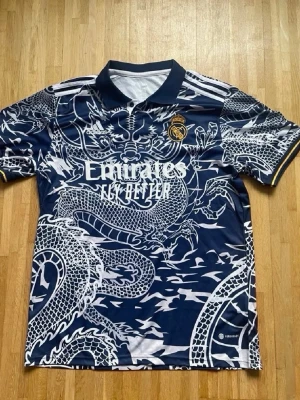 Real Madrid draktröja Adidas - Snygg mörkblå Real Madrid-tröja från Adidas med ett stort vitt drakmönster över hela plagget. Tröjan har korta ärmar, krage och klubbens emblem på bröstet. Materialet är lätt och andas, perfekt för sport och träning.
