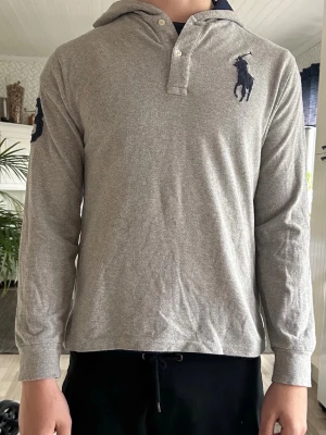 Grå hoodie från Polo Ralph Lauren - Säljer en grå hoodie från Polo Ralph Lauren med klassisk logga broderad på bröstet och ärmen. Hoodien har lång ärm, knappar vid halsen och är gjord i mjukt bomullsmaterial. Perfekt för dig som gillar stilren design och ikoniska märken.