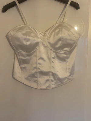 Vit satin korsettopp med rosett - Vit korsettopp i glansigt satinmaterial med tunna axelband och markerade kupor. Toppen har en liten söt rosett framtill och justerbara axelband. Jättefin att ha både på utekväll eller till vardags. Aldrig använd💕💕