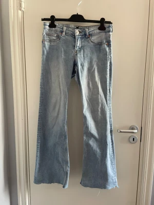 Ljusblå bootcut jeans med rå kant - Säljer ett par ljusblå bootcut jeans med rå, fransig kant längst ner. Jeansen har klassiska fem fickor och knappgylf. Perfekt för dig som gillar en avslappnad och trendig look. Materialet är denim.