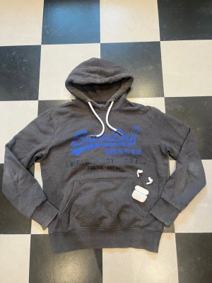 Grå Superdry hoodie med blå logga - Snygg grå hoodie från Superdry med stor blå logga och text på bröstet. Klassisk känguruficka framtill och vita dragsnören i huvan. Mjuk bomullsmix som känns skön mot huden. Perfekt för en avslappnad streetwear-look.