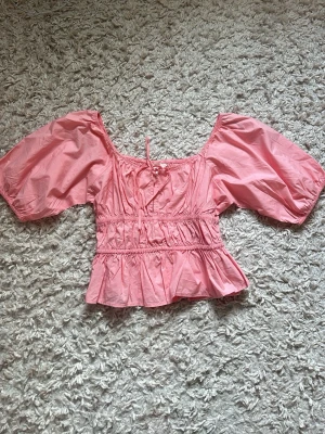 Rosa blus med puffärmar - Säljer en söt rosa blus med puffiga korta ärmar och smockad midja. Blusen har en volang nertill och en liten knytning vid halsen. 🩷🩷🩷