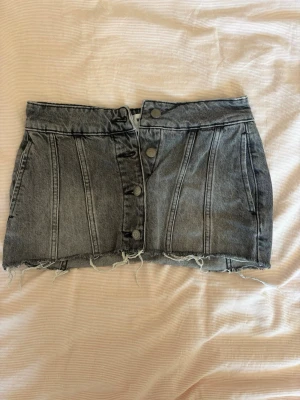 Grå denim kortkjol med knappar - Cool grå denimkjol från Weekday med rå, fransig nederkant och dekorativa sömmar. Kjolen har knappar framtill och är endast använd en gång. Storlek 34