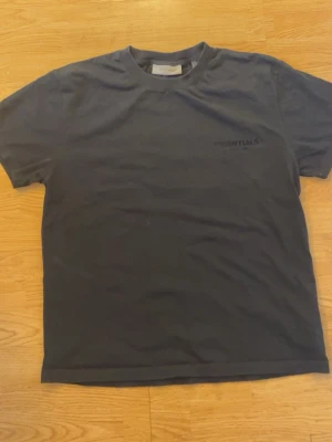 Svart Essentials Fear of God t-shirt - Säljer en svart t-shirt från Fear of God Essentials med diskret logga framtill och stor ESSENTIALS-text på ryggen. Klassisk passform med rund hals och korta ärmar. Tillverkad i mjuk bomull som känns skön mot huden.