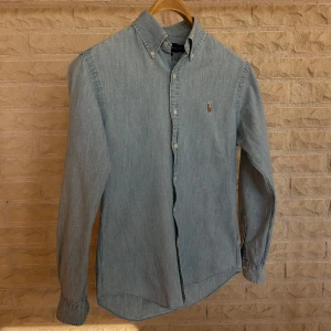 Ljusblå jeansskjorta med knappkrage - Säljer en stilren ljusblå skjorta med knappkrage. Skjortan har långa ärmar och en liten broderad detalj på bröstet. Perfekt för både vardag och fest! 🕺