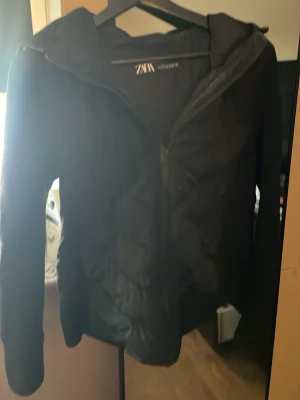 Svart slimthick jacka  från Zara - Säljer en svart pufferjacka från Zara med huva och dragkedja framtill. Jackan är tillverkad i ett slätt syntetmaterial och har en enkel, stilren design som passar perfekt för kalla vinterdagar. Perfekt för dig som vill ha en varm och snygg jacka.
