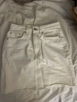 Vit jeanskjol med rå kant - Säljer en vit kort jeanskjol med fem fickor och rå, fransad nederkant. Kjolen har knapp och dragkedja framtill samt bälteshällor. Perfekt för dig som gillar en enkel men trendig stil.