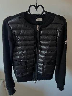 Svart cardiganjacka från Moncler - Snygg svart cardiganjacka från Moncler med två fickor med knappstängning. Jackan har rund halsringning, ribbade muddar och dragkedja framtill. Mycket fint skick!