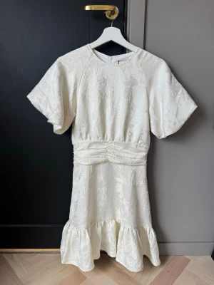 Vit kortklänning med volang - Supersöt vit kortklänning med strukturmönster och volang nedtill. Klänningen har korta ärmar, markerad midja med rynkdetaljer och ett diskret blommigt mönster i tyget. Perfekt för studenten! Aldrig använd, helt ny! ☺️