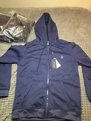 Marinblå och svart hoodie från Polo Ralph Lauren - Två snygga hoodies från Polo Ralph Lauren i marinblått och svart. Klassisk design med dragkedja, huva med dragsko och fickor framtill. Diskret logga broderad på bröstet. Tillverkad i mjukt bomullsmaterial som känns skönt mot huden.