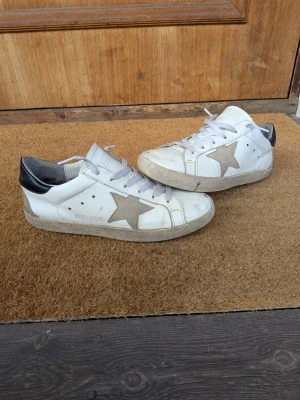 Golden Goose vita sneakers med stjärna - Golden Goose sneakers i vitt läder med grå stjärna på sidan och svart detalj på hälen. Klassisk låg modell med vita skosnören och perforeringar vid tån. Snygg och trendig design som passar till många outfits.
