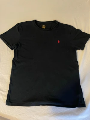 Svart t-shirt från Polo Ralph Lauren - Klassisk svart t-shirt från Polo Ralph Lauren med röd broderad logga på bröstet. T-shirten har rund halsringning och korta ärmar. Perfekt basplagg som funkar till allt och är tillverkad i mjuk bomull.
