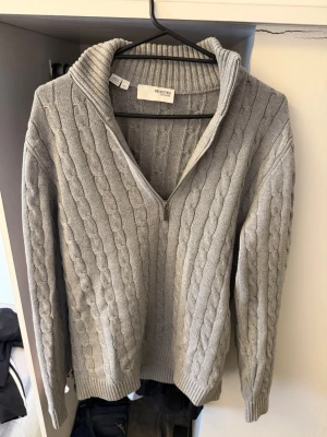 Grå kabelstickad tröja Selected Homme - Kabelstickad grå tröja från Selected Homme med halv dragkedja och hög krage. Tröjan har långa ärmar, ribbade muddar och är tillverkad i mjukt stickat material. Perfekt för lager-på-lager och en stilren look.