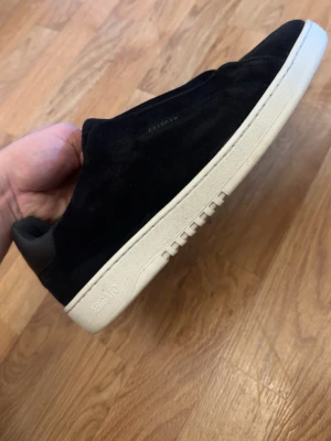 Svarta Arigato sneakers laceless - Stilrena svarta sneakers från Axel Arigato i mjuk mocka med vit sula. Diskret logga på sidan och elastisk detalj över vristen för en clean look. Perfekta för dig som gillar minimalistisk design och premiumkänsla.