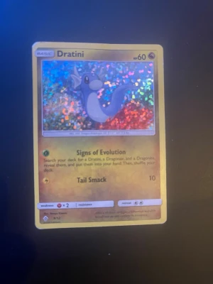 Holo Dratini Pokémonkort - Säljer ett holografiskt Dratini-kort från Pokémon. Kortet har en skimrande bakgrund med färgglada effekter och visar Dratini i tecknad stil. Text och detaljer är på engelska. Perfekt för samlare eller Pokémonfans!