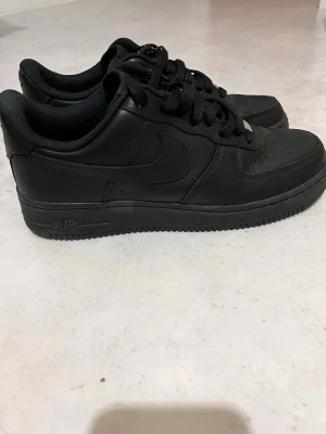 Svarta Nike Air Force 1 sneakers - Klassiska helsvarta Nike Air Force 1 sneakers i lågt utförande. Skorna är tillverkade i slätt läder med matchande svarta skosnören och sula. Ikonisk Nike Swoosh på sidan och AIR-detalj på sulan. Perfekta för en clean och stilren look. Kan också diskutera om priset 