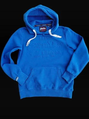Superdry hoodie - Blå hoodie från Superdry med broderad logga på bröstet och vita dragsnören i huvan. Storlek S men liten i storlekenKlassisk känguruficka framtill och ribbade muddar vid ärmar och nederkant. Perfekt för en avslappnad och cool stil. Hör av er om ni har frågor!