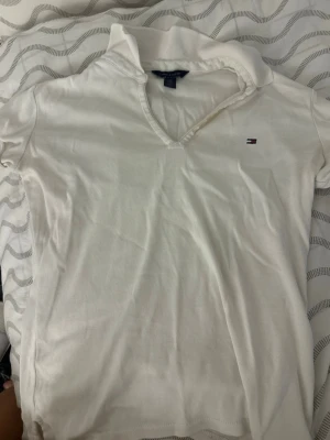 Vit pikétopp från Tommy Hilfiger - Klassisk vit pikétopp från Tommy Hilfiger med liten broderad logga på bröstet. Toppen har korta ärmar, v-ringning och krage. Tillverkad i mjuk bomull som känns skön mot huden. Perfekt till jeans eller kjol för en clean och enkel look.