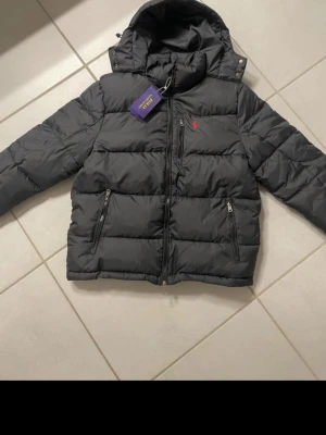Svart pufferjacka från Polo Ralph Lauren - Snygg svart pufferjacka från Polo Ralph Lauren med röd broderad logga på bröstet. Jackan har huva, två dragkedjeförsedda fickor och quiltad design. Perfekt för kalla dagar och riktigt trendig look. Hör av er vid funderingar 