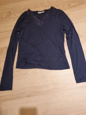 Mörkblå långärmad topp med spets - Mörkblå långärmad topp med v-ringning och dekorativ spetskant runt halsen. Tunn och mjuk i materialet. Enkel och stilren design som passar till mycket.