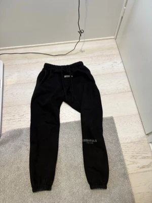 Svarta Essentials mjukisbyxor - Säljer ett par svarta mjukisbyxor från Essentials med tryckt logga på vänster ben. Byxorna har resår i midjan och vid bensluten, samt fickor på sidorna och en bakficka. Perfekta för chill dagar och streetwear-stil. There is a small stich in right knee because there was a hole
