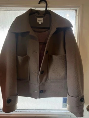 Beige ulljacka från Kappahl - Stilren beige jacka från Kappahl i ullmix med stora fickor framtill, bred krage och knappar. Jackan har en boxig passform och uppvikta ärmslut med dekorativa knappar. Perfekt för dig som gillar minimalistisk och trendig stil. Perfekt för dig som är runt 14 år! En söm har gått upp lite på ena armen men inget som märks eller syns när man har jackan på.🌸