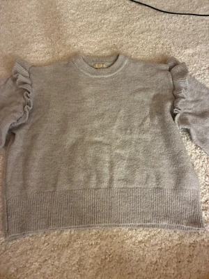 Grå stickad tröja med volangärmar - Supersnygg grå stickad tröja från Gina Tricot med volangdetaljer längs ärmarna. Tröjan har rund halsringning, ribbstickade muddar och är gjord i mjukt material. Perfekt för dig som vill ha en trendig och bekväm look.