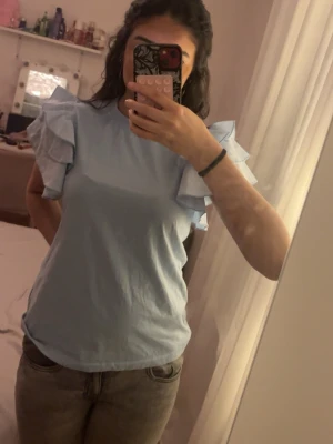 Ljusblå topp med volangärmar - Supersöt ljusblå topp med volangdetaljer på ärmarna. T-shirten har rund halsringning och är tillverkad i mjukt bomullstyg. Perfekt för dig som vill ha en enkel men ändå unik look med extra fokus på axlarna.