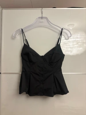 Svart korsettopp med knappar från Zara  - Snygg svart korsettopp med tunna axelband och knappar framtill. Toppen har markerade kupor och lätt utsvängd nederdel för en feminin siluett. Perfekt att styla med jeans eller kjol för en trendig look. I storlek xs från Zara 