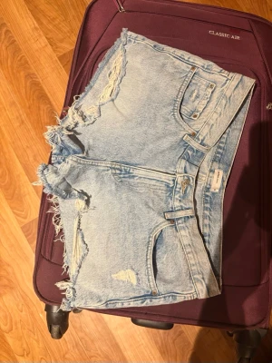 Ljusa jeansshorts med fransar - Säljer ett par ljusblå jeansshorts från Gina Tricot med slitna detaljer och fransiga kanter. Klassisk femficksmodell med råa benslut och hög midja. Perfekta för sommaren och ger en avslappnad vibe.