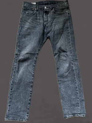 Levis 501 - Säljer ett par grå jeans från Levi's med klassisk rak passform och fem fickor. Jeansen har en diskret tvättad look och är tillverkade i slitstark denim. Perfekta för dig som gillar en enkel och tidlös stil.