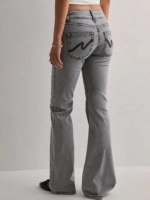Grå bootcut jeans från Nelly - Säljer ett par grå bootcut jeans från Nelly med klassiska kontrastsömmar och broderade bakfickor. Jeansen har låg midja och utsvängda ben, perfekta för en avslappnad och trendig look. Jeansen är lite trasiga vid benen men det går att klippa annars är de rena. Pris kan diskuteras!!