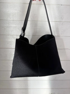 Svart axelremsväska  ginatricot - Nypris: 500kr Stilren svart handväska med axelrem i läder och kropp i mjuk mocka. Väskan har en unik asymmetrisk form och stängs med magnetknapp. Insidan är fodrad i svart läder och har en metallspänne på remmen för extra detalj.