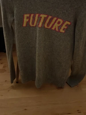 From Future Tröja - Stickad tröja i mjuk grå ton med texten FUTURE i stora bokstäver framtill i gult och rosa. Tröjan har långa ärmar och ribbade muddar vid ärmslut och nederkant. Perfekt för dig som gillar trendiga statement-plagg.
