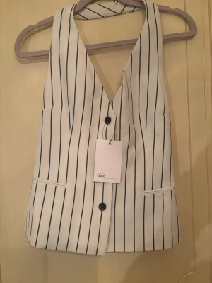 Vit randig väst från ASOS - Snygg vit väst från ASOS med svarta vertikala ränder och två knappar framtill. Västen har v-ringning och två fickor, perfekt för att lyfta din outfit med en stilren touch.