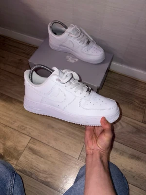 Nike Air Force 1 vita sneakers - Klassiska Nike Air Force 1 sneakers i helvitt läder med vit sula och vita skosnören. Ikonisk design med perforerad tåbox och Nike swoosh på sidan. Passar perfekt till streetwear och har en tidlös look.