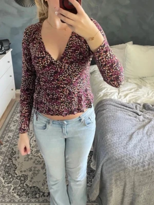 Blommig omlottopp med v-ringning - Säljer en blommig omlottopp i mörka toner med v-ringning och långa ärmar. Toppen har en figurnära passform och är tillverkad i ett mjukt, stretchigt material. Perfekt att styla med jeans eller kjol för en trendig look.