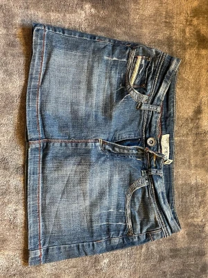 Blå jeanskjol från D*KLIC - Säljer en klassisk blå jeanskjol från D*KLIC med fem fickor och orangea kontrastsömmar. Kjolen har en kort modell och stängs med knapp och dragkedja framtill. Motsvarar storlek 36