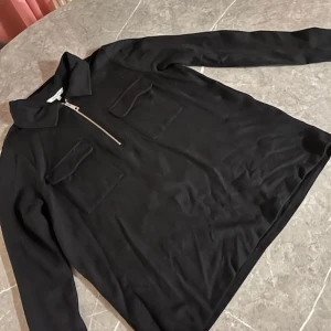 Svart långärmad tröja med dragkedja - Svart långärmad tröja med krage och halv dragkedja framtill. Två bröstfickor med lock ger en cool utility-vibe. Perfekt till jeans eller kjol för en avslappnad men snygg look. Materialet ser ut att vara mjukt och stretchigt.