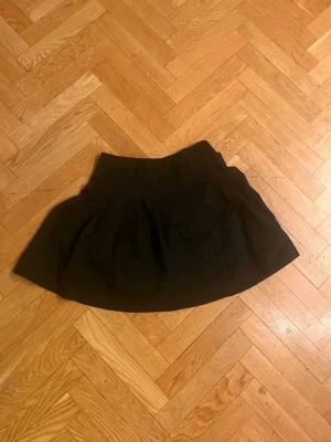Svart vid kortkjol - Säljer en svart kortkjol med vid passform och fina veck. Kjolen har en enkel, stilren design och är perfekt för dig som gillar en klassisk look. Materialet känns som bomull eller polyester och kjolen har en hög midja. Skriv gärna för mått! 💕