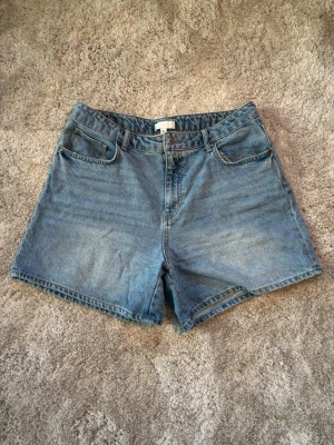 Blå denimshorts med fem fickor - Klassiska blå jeansshorts i bomull med fem fickor och knappgylf. Modellen har en rak passform. Perfekta för sommaren och enkla att matcha med olika stilar.