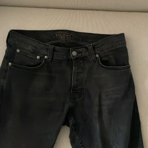 Nudie Jeans Grim tim - Säljer ett par svarta jeansbyxor från Nudie Jeans med klassisk femficksdesign och normal passform. Byxorna har dragkedja och knapp i midjan samt raka ben. Perfekta för en stilren look.