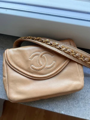 Äkta Chanel väska (vintage) - belt bag / bum bag  - Säljer en beige handväska från Chanel i mjukt läder med klassiskt CC-logo broderat på locket. Väskan har en axelrem med guldfärgad kedjedetalj invävd i läder. Perfekt storlek för det viktigaste och en tidlös design.