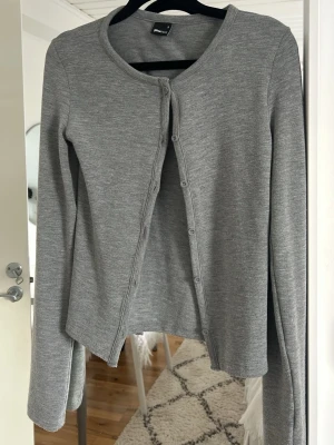 Grå kofta från Gina Tricot - Snygg grå kofta från Gina Tricot med rund halsringning och knäppning framtill. Tillverkad i mjukt, ribbat material och har långa ärmar. Perfekt till sommaren 