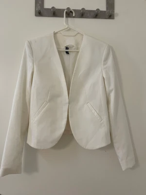 Vit kavaj från H&M - Stilren vit kavaj från H&M med öppen front och två dekorativa fickor. Kavajen har lång ärm och en något figurnära passform. Perfekt för att lyfta vilken outfit som helst med en fräsch och modern look.