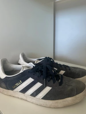 Adidas Gazelle mörkblå sneakers - Säljer ett par klassiska Adidas Gazelle sneakers i mörkblå mocka med vita detaljer. Skorna har vita sulor och snörning framtill. Det är använda en del så färgen är lite urtvättad. Perfekta för dig som gillar retrostil och streetwear.