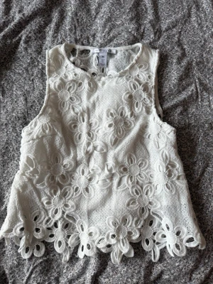 Vit spetsblus från NLY Trend - Säljer en superfin vit blus från NLY Trend i storlek 36. Toppen är ärmlös och har ett blommigt spetsmönster med snygga detaljer längs kanten. Perfekt till sommaren och ger en romantisk vibe till din outfit.